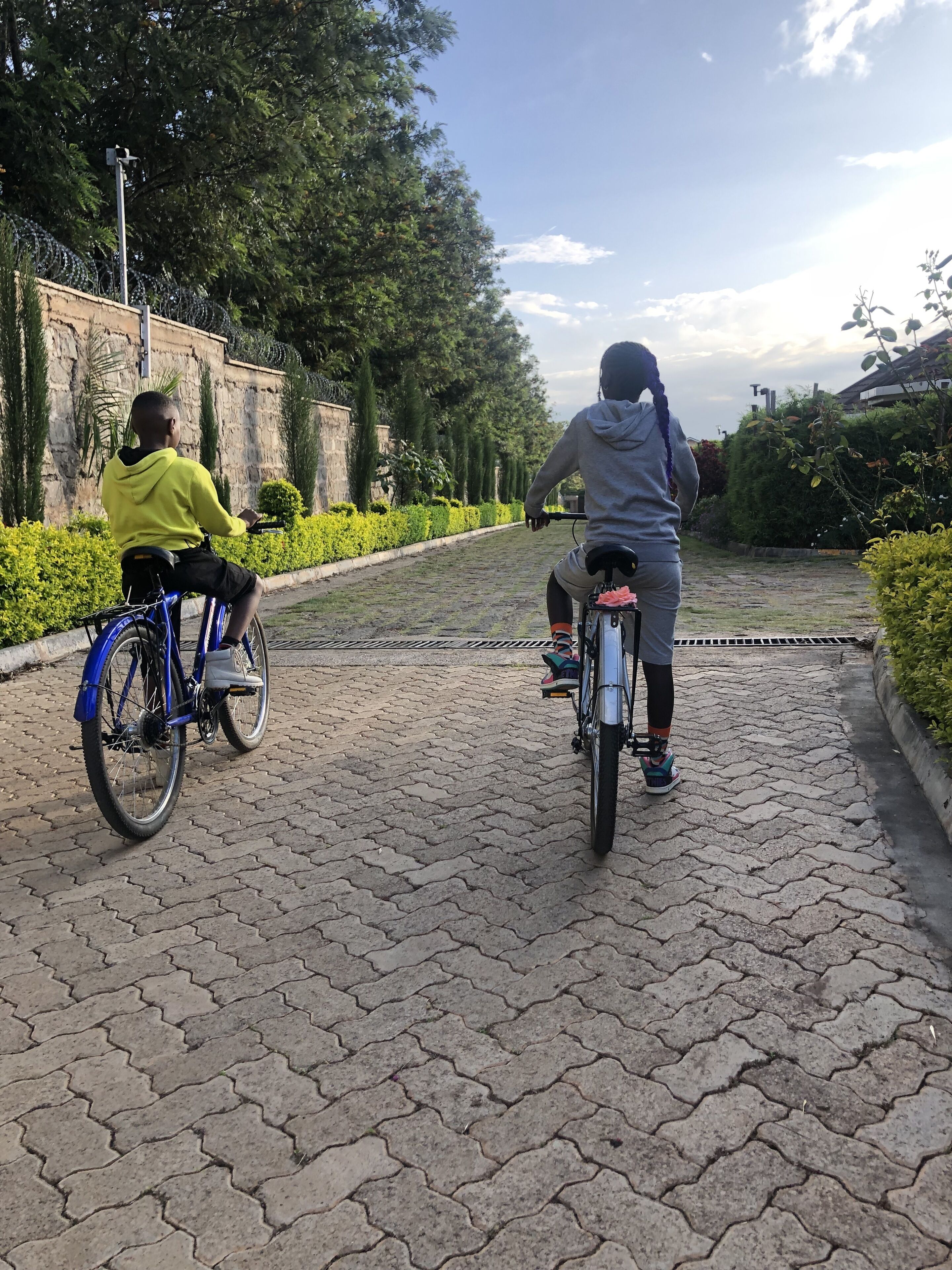 Two people riding bicycles on a paved pathway surrounded by greenery and a stone wall.
