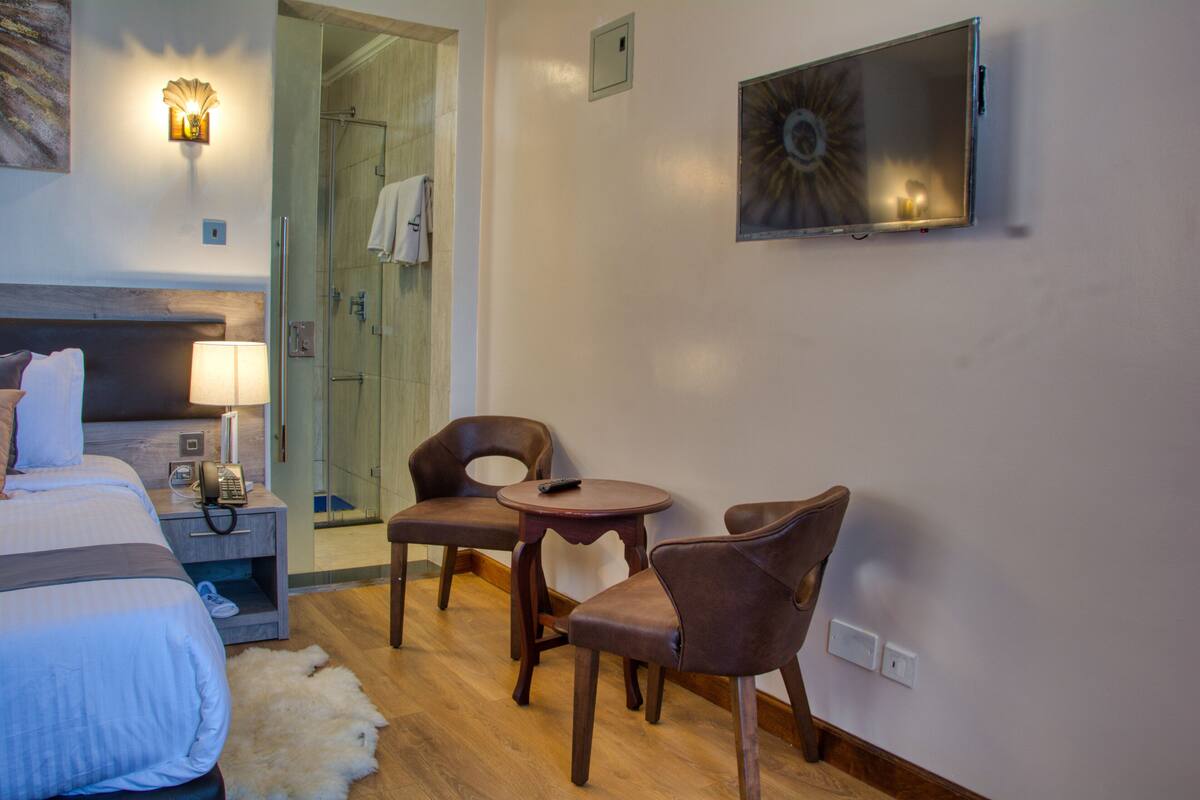 Habitación de hotel con cama, mesita de noche con lámpara, teléfono, dos sillas, una mesita pequeña, televisor montado en la pared y zona de ducha visible a través de una puerta abierta.