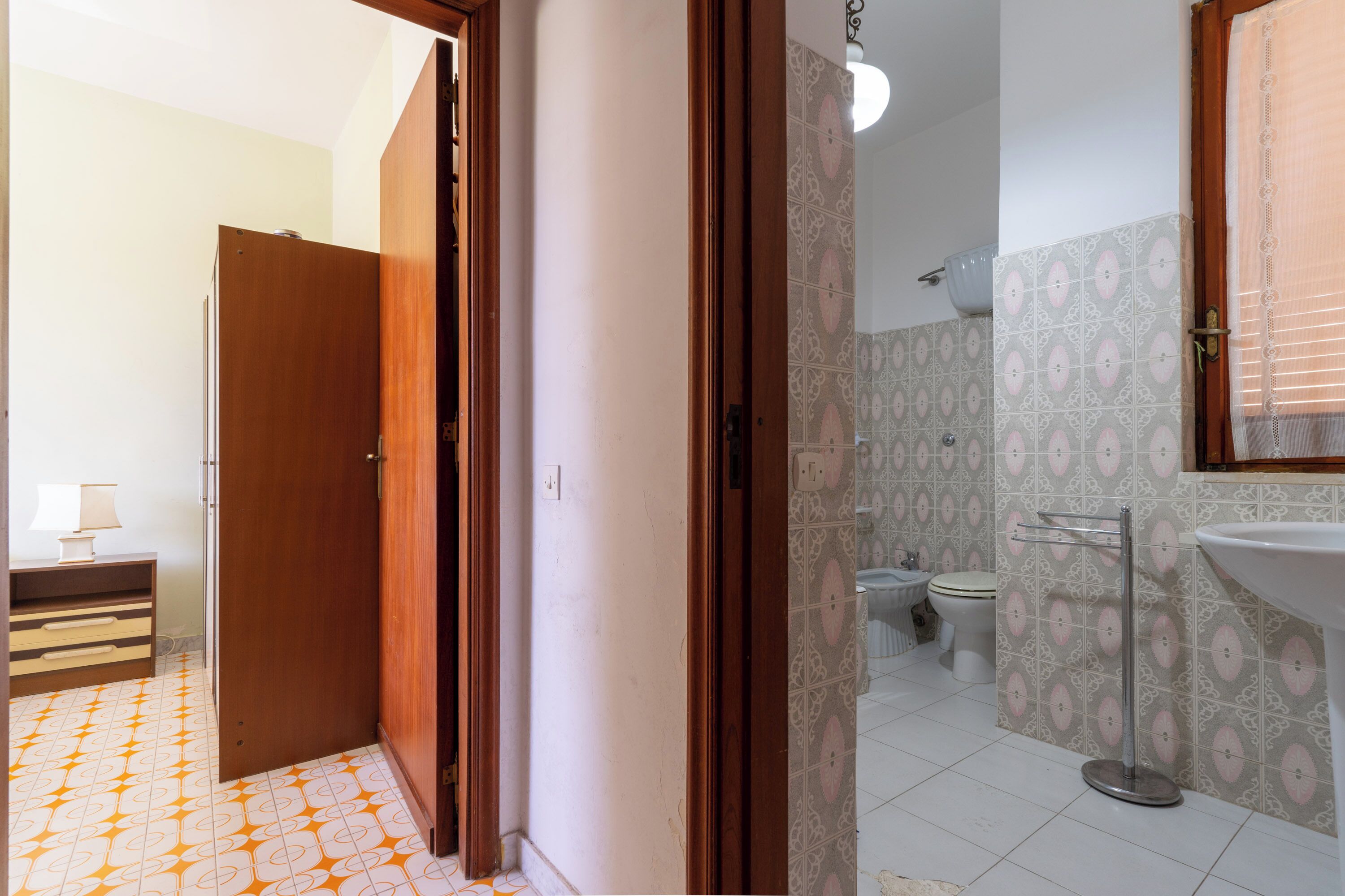 A bathroom with a toilet, bidet, and sink. There is a wooden door leading to another room. The floor has a patterned tile design.