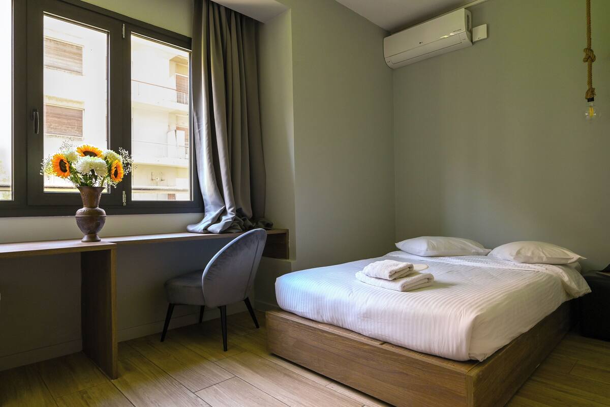A bedroom with a bed, a desk, a chair, a window with curtains, and an air conditioning unit.