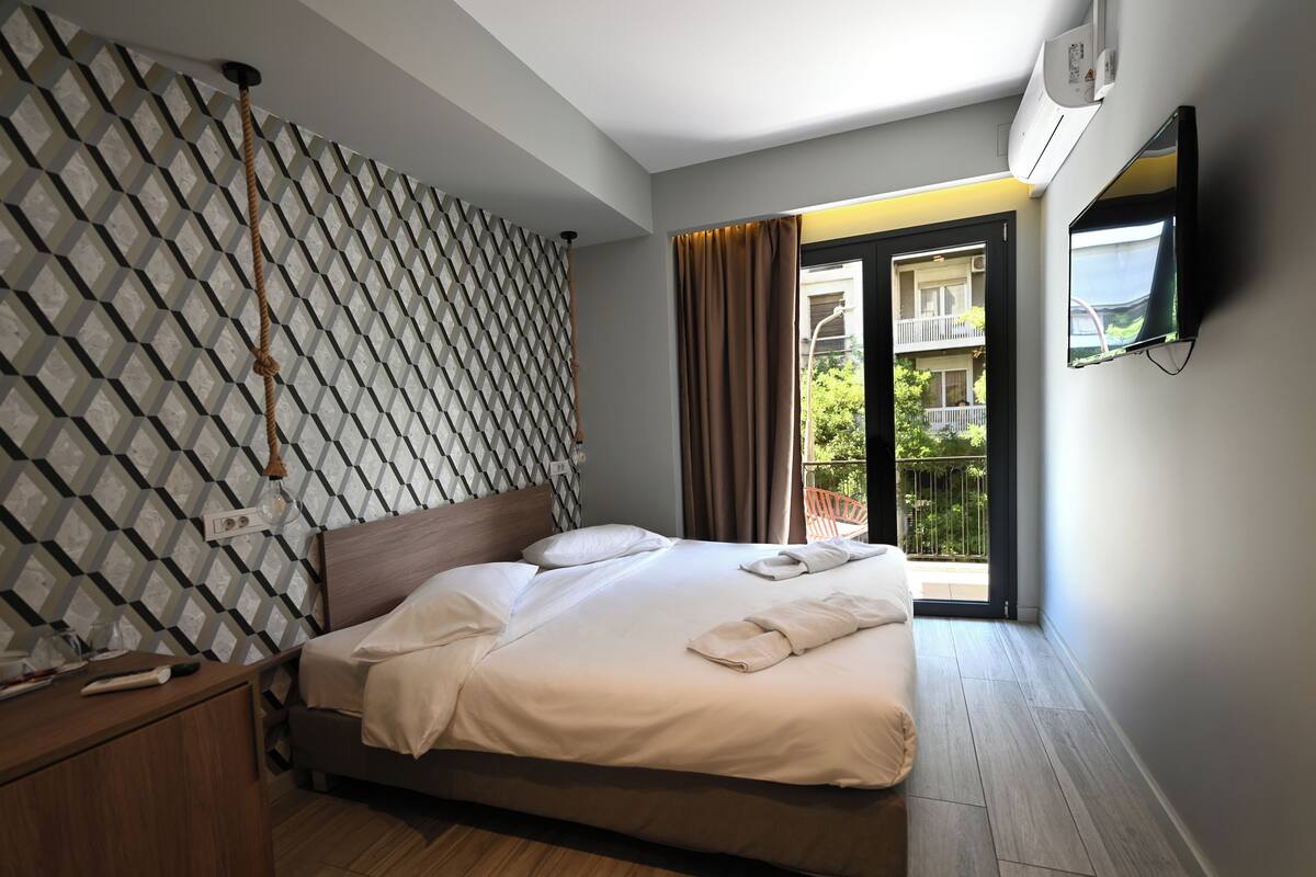 A modern hotel room with a patterned wall, a wooden headboard, a neatly made bed, a TV, and a balcony with a view.