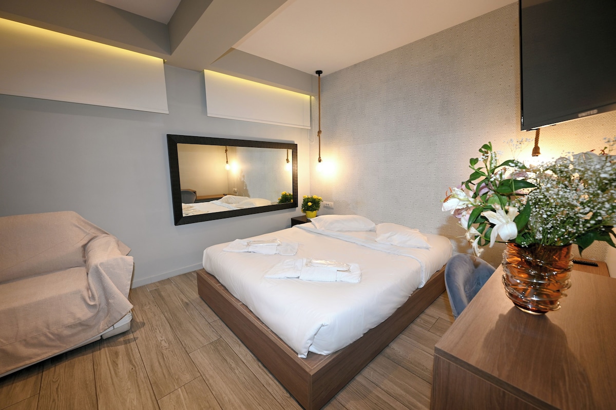 Ein modernes Hotelzimmer mit einem großen Bett, einer Couch, einem Nachttisch aus Holz, einem Spiegel und einer Blumenvase.