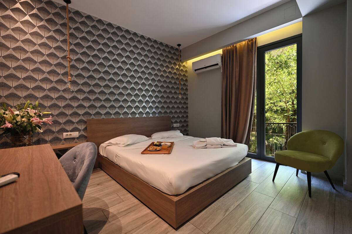 A modern hotel room with a geometric-patterned wall, a bed with white linens, a wooden desk, a green armchair, and a balcony with a view of greenery.