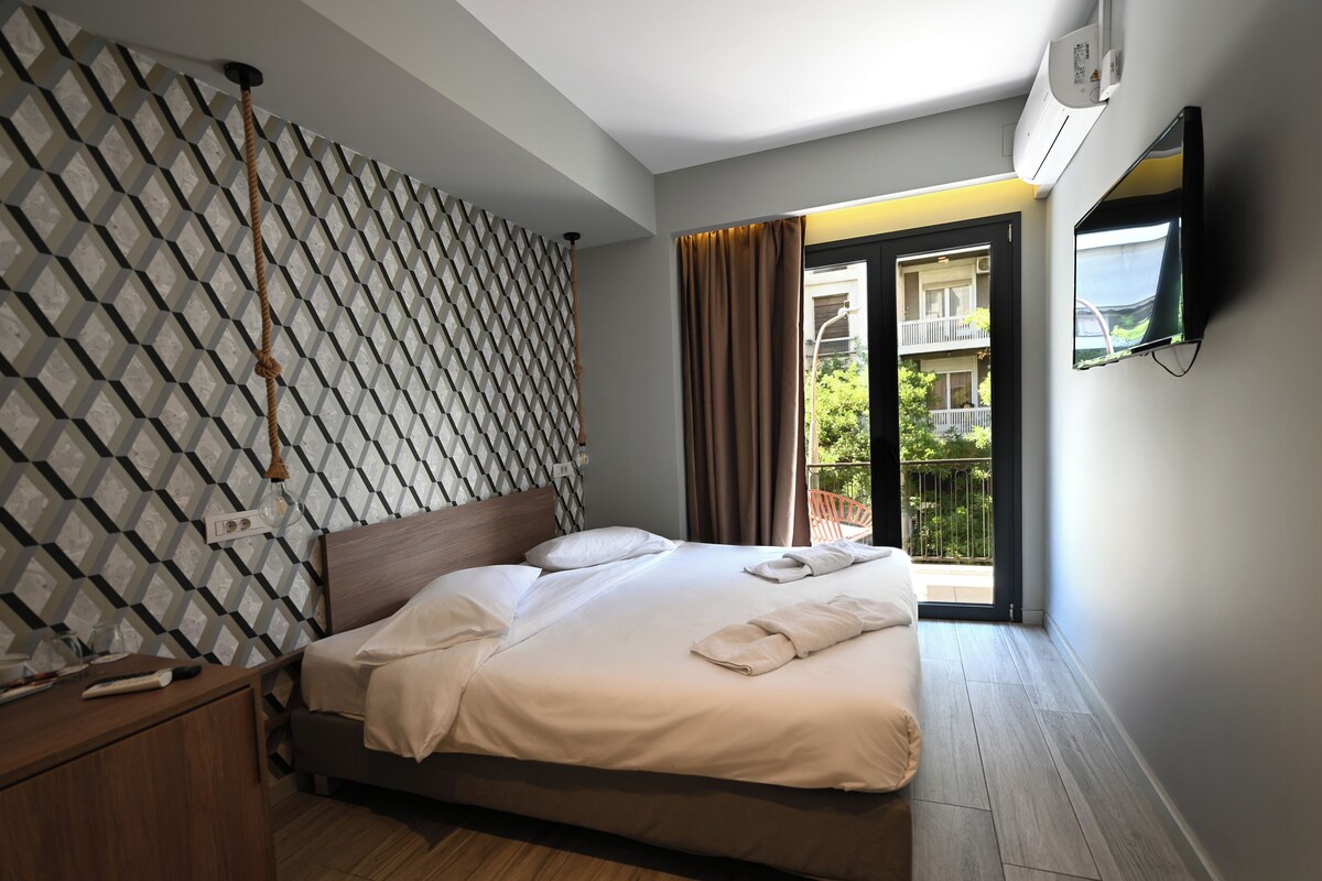 Camera d'albergo moderna con parete decorata, una testiera in legno, un letto rifatto e una televisione, con un balcone e vista panoramica.