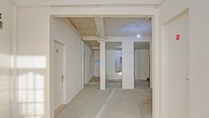 Un couloir aux murs et aux portes blancs, certaines portes étant signalées par des panneaux rouges, et dont le sol est en béton.