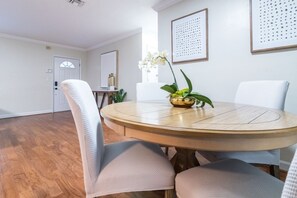 A dining area with a round wooden table, white upholstered chairs, and a potted plant.