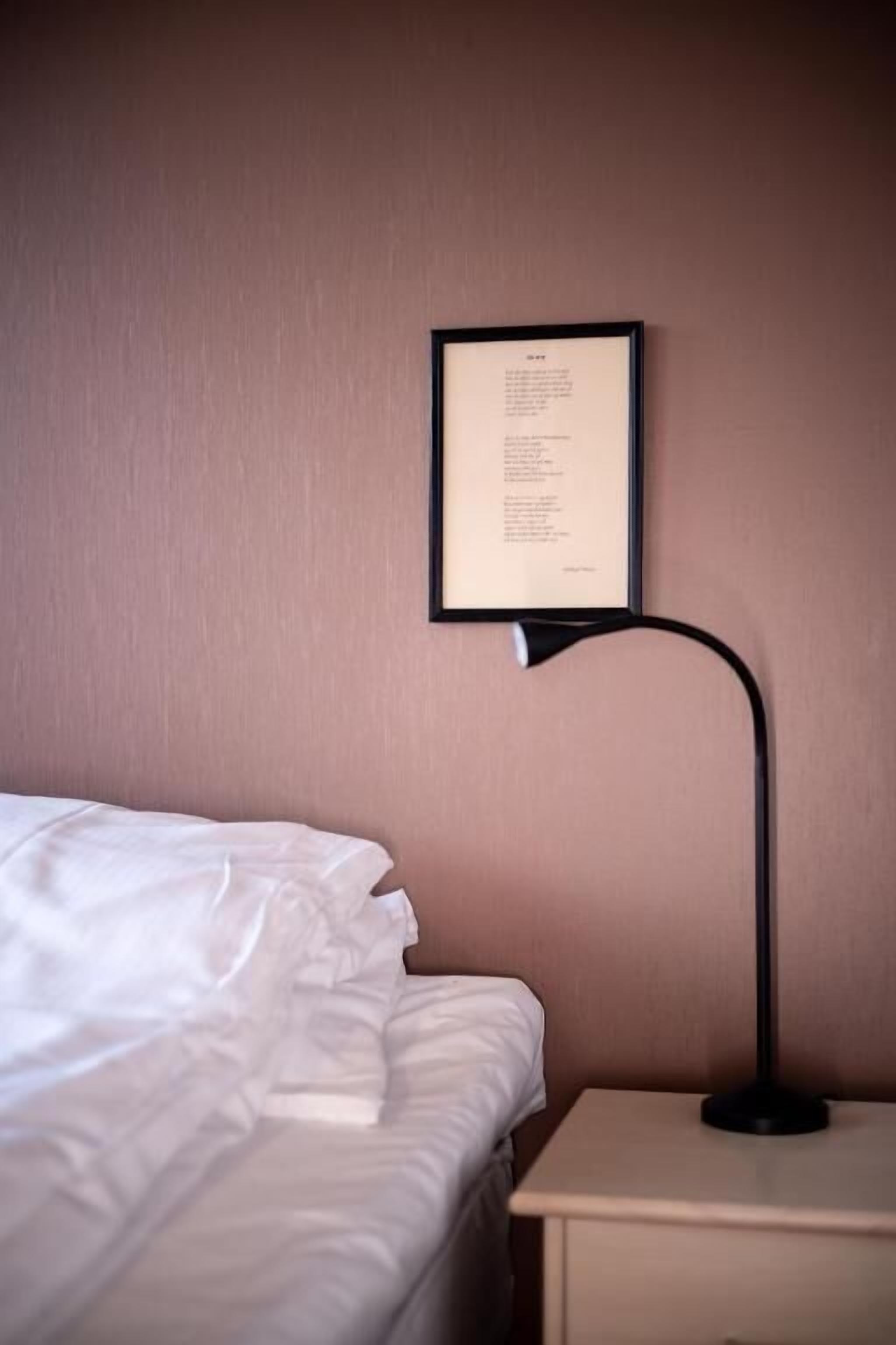 Una cama bien hecha con sábanas blancas, una mesita de noche con una lámpara y un texto enmarcado en la pared.