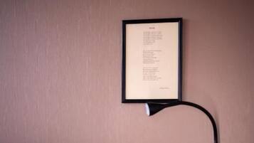 Una cama bien hecha con sábanas blancas, una mesita de noche con una lámpara y un texto enmarcado en la pared.
