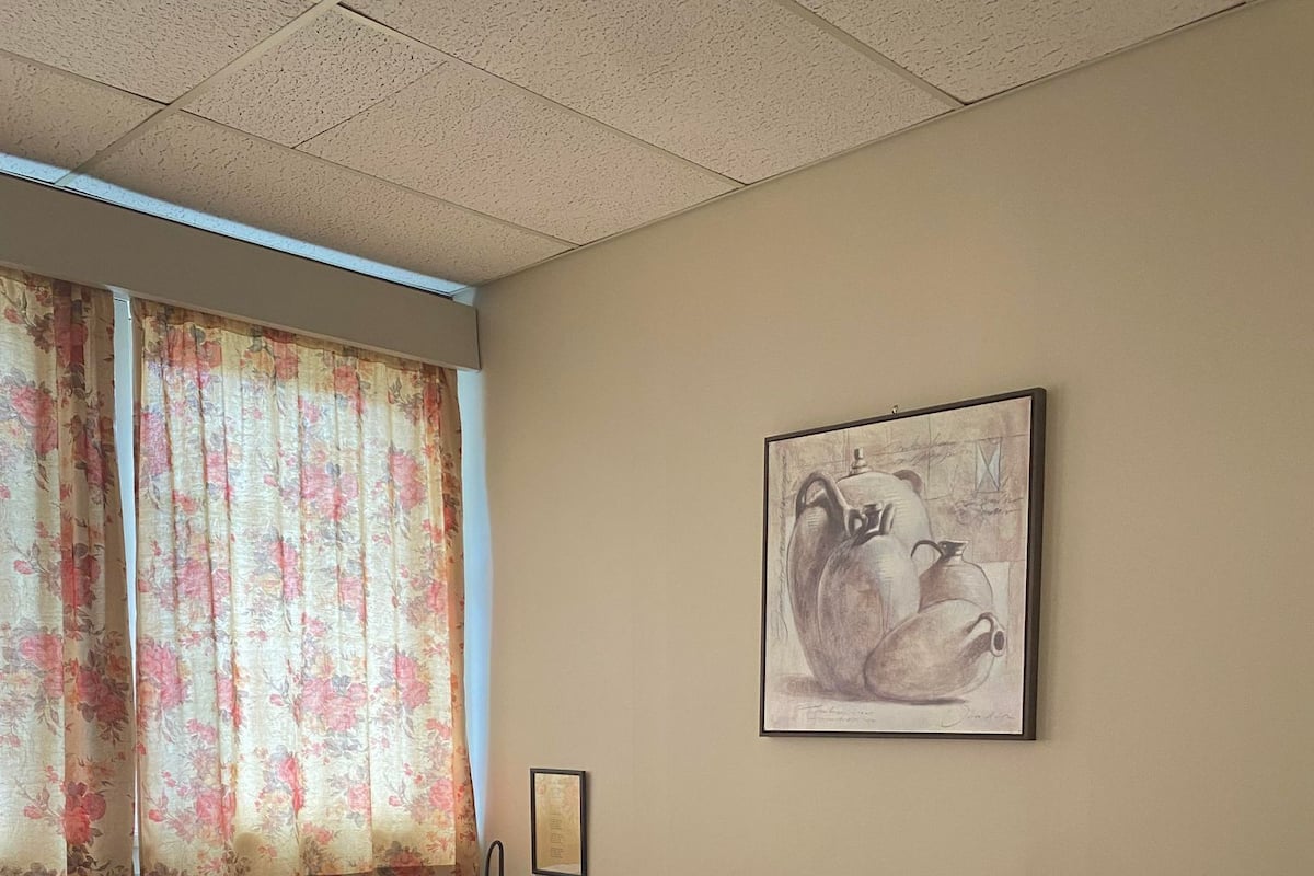 Ein Hotelzimmer mit zwei Betten, einem Fenster mit floralen Vorhängen, einem gerahmten Kunstwerk an der Wand und einem Nachttisch mit Lampe.
