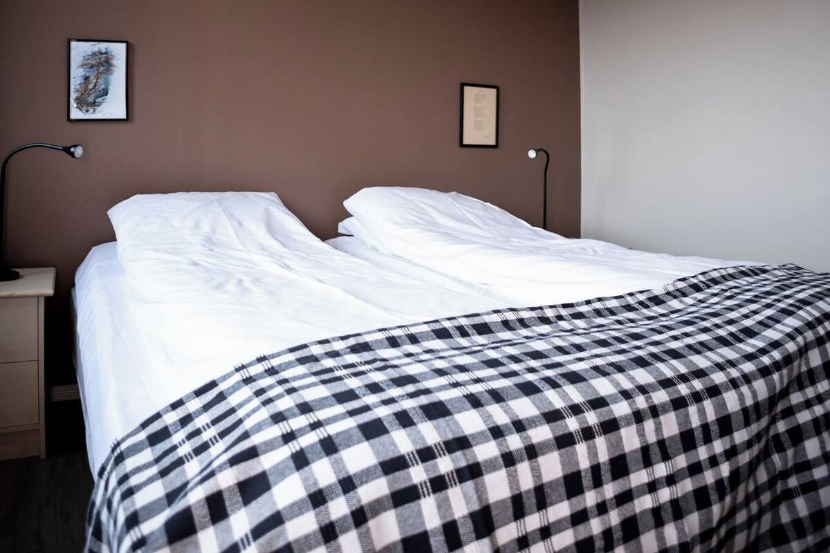 Ein ordentlich bezogenes Bett mit weißen Leinen und einer karierten Decke, eine Nachttischlampe und ein gerahmtes Bild an der Wand.