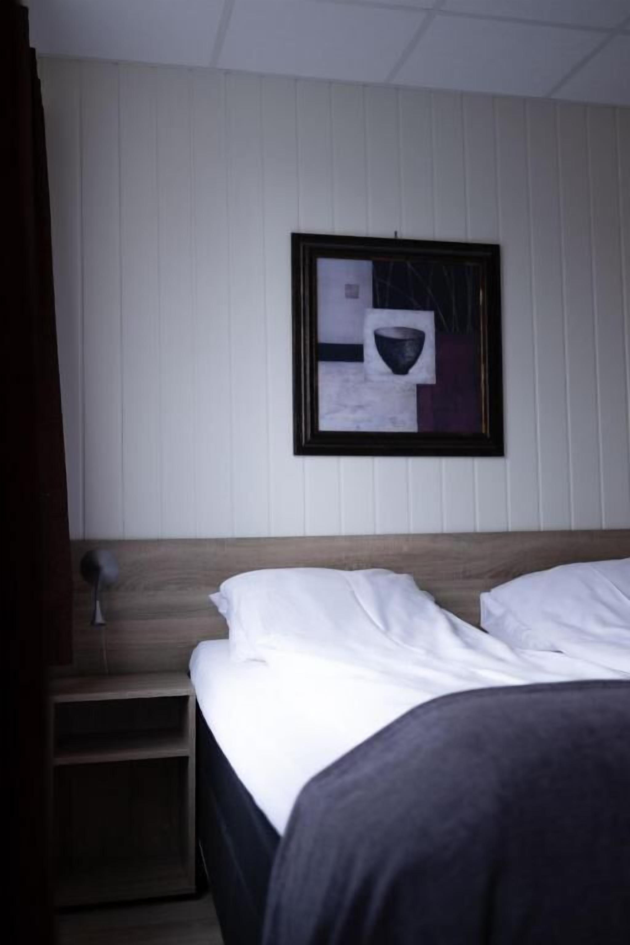 A hotel room with a wooden headboard, a bed with white linens, a nightstand with a lamp, and a framed picture on the wall.