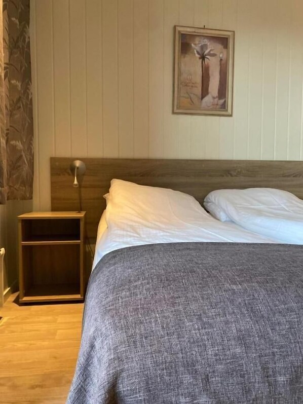 Una cama bien hecha con ropa de cama blanca y gris, una mesita de noche de madera y un cuadro en la pared.