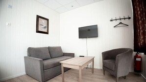 Una sala de estar moderna con un sofá gris, una mesa de centro de madera y un sillón gris. Un televisor de pantalla plana está montado en la pared, y se observa un extintor a la derecha.