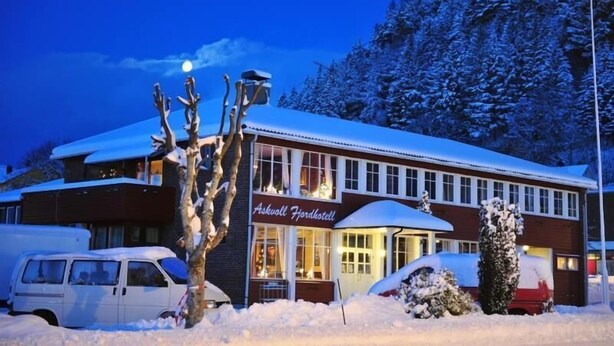 Un hotel con fachada nevada, entrada visible y vehículos estacionados al frente.