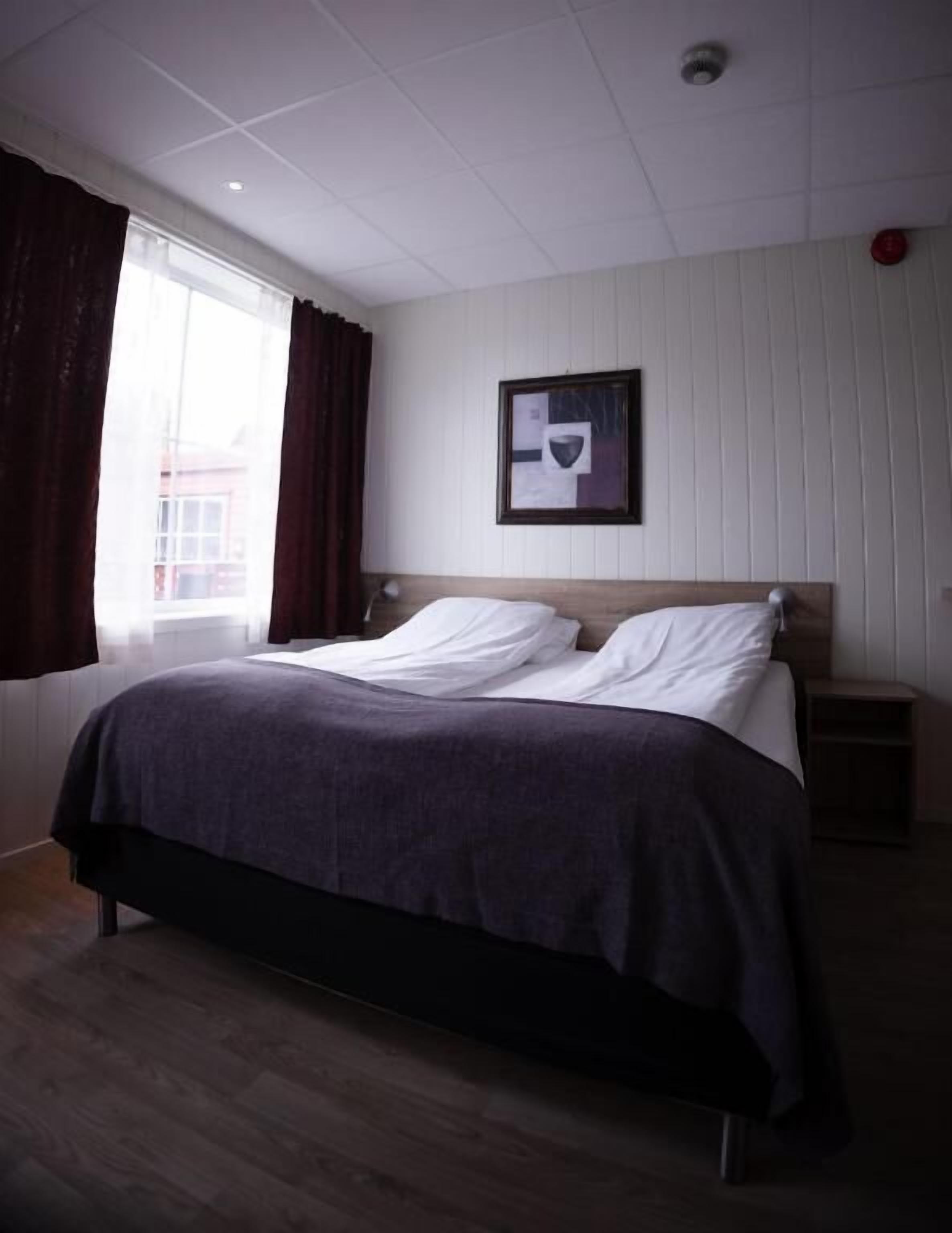 Un dormitorio con una cama, una mesita de noche, una ventana con cortinas e imagen colgada en la pared.