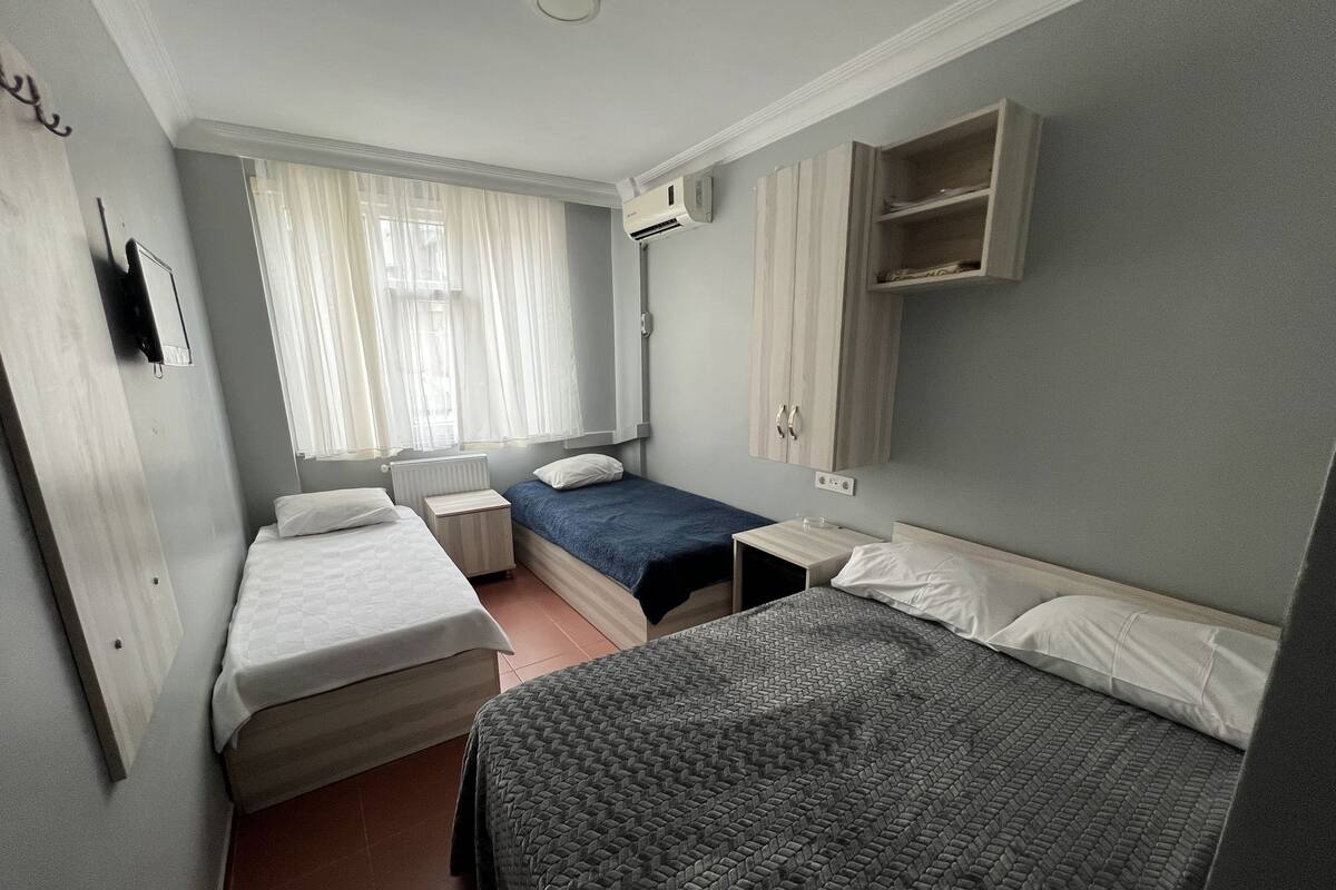 Una habitación pequeña y luminosa con dos camas individuales, un armario de madera, una mesita de noche y una ventana con cortinas translúcidas.