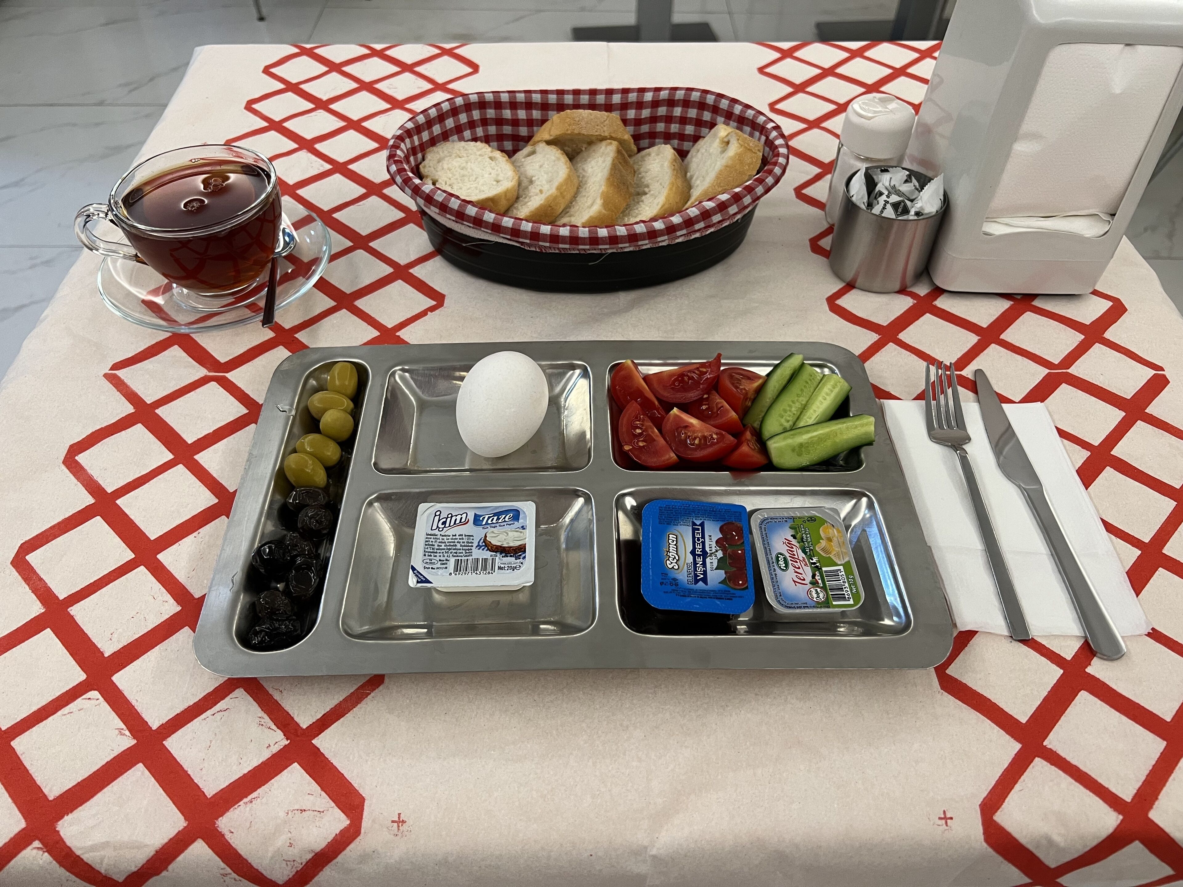A meal tray with bread, eggs, tomatoes, cucumbers, olives, and condiments on a table with a checkered tablecloth.