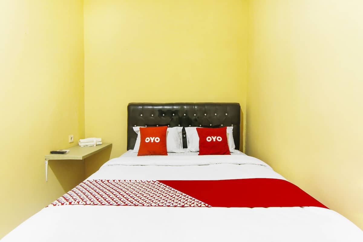 Quarto de hotel com cama, duas almofadas vermelhas com a inscrição "OYO", mesa de cabeceira com telefone e colcha de cama com padrão vermelho e branco.