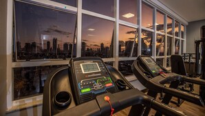 Ein Fitnessstudio mit Laufbändern, großen Fenstern und Blick auf die Stadt bei Sonnenuntergang.