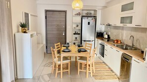 A modern kitchen with white cabinets, a built-in oven, a refrigerator, and a dining area with a wooden table and chairs. - GPK Lux 2BR | 7 min walking to Acropolis (Athina)