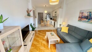 A modern living room with a grey sofa, wooden herringbone flooring, a white coffee table, and a dining area with a wooden table and chairs. - GPK Lux 2BR | 7 min walking to Acropolis (Athina)