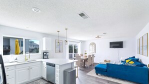 A modern kitchen with white cabinetry, a central island, and a dining area with a blue sofa and a TV.