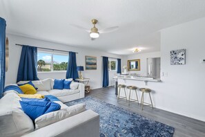 Sala moderna com sofá branco, almofadas azuis e amarelas, tapete azul e um espaço de jantar com banquetas.
