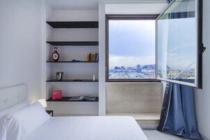 Habitación de hotel con cama, estantería, ventana con vista a un paisaje urbano y cortina.