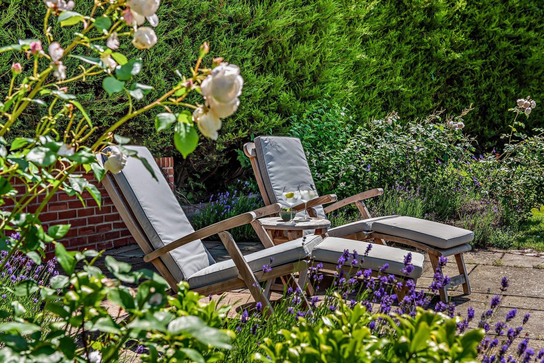 Ein Garten mit Sitzgelegenheit, bestehend aus zwei Holzliegen mit Kissen, einem kleinen runden Tisch und einer Getränkflasche.