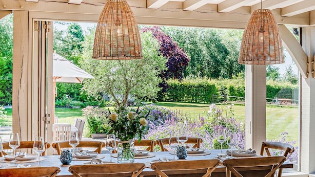 An outdoor dining area with wooden tables and chairs, a wicker pendant light, and a view of a garden with purple flowers and a lawn.