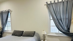 Ein Schlafzimmer mit einem Bett, zwei Fenstern mit Vorhängen, einem Nachttisch mit Lampe und Blick ins Freie.