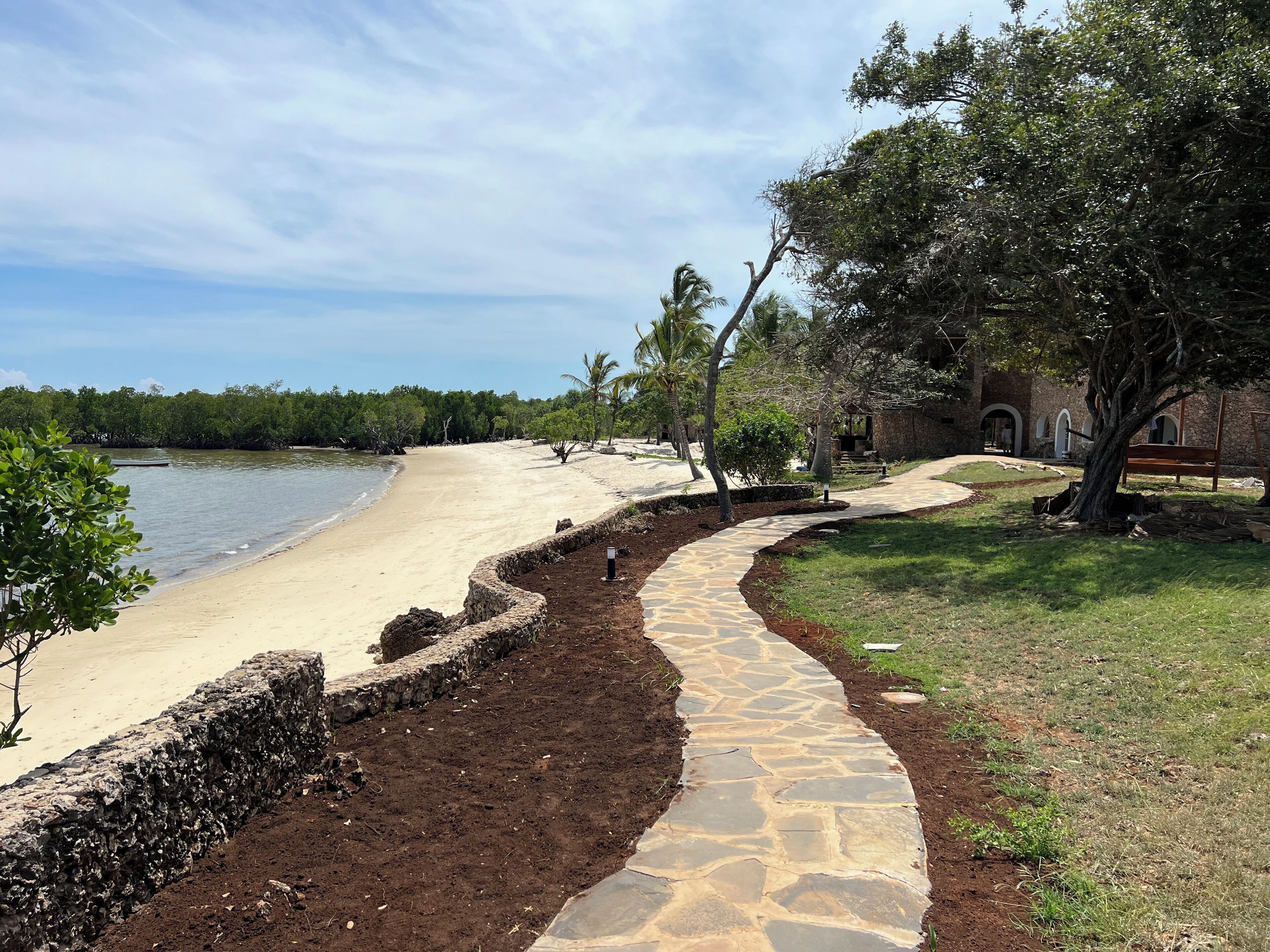 Un sentier côtier qui mène à une plage, avec un mur de pierres et une zone herbeuse bordée d’arbres.