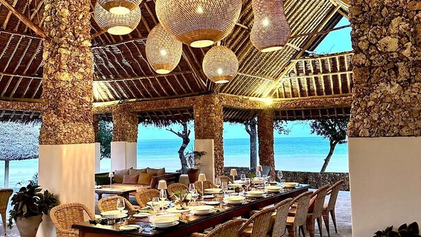A dining area with a long table set for a meal, surrounded by wicker chairs, under a thatched roof with hanging spherical light fixtures. - Mwazaro Beach Lodge (Shimoni)