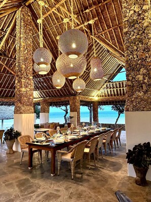 A dining area with a long table set for a meal, surrounded by wicker chairs, under a thatched roof with hanging spherical light fixtures. - Mwazaro Beach Lodge (Shimoni)