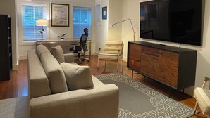 Ein modernes Wohnzimmer mit einem Ecksofa, einem Flachbildfernseher, einer Holzkommode und einem Schreibtisch mit Stuhl.