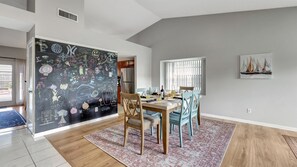 Ein Essbereich mit einem Holztisch, Stühlen und einem gemusterten Teppich. Eine Wand mit Kreidetafel zeigt verschiedene Zeichnungen und Schriftzüge.