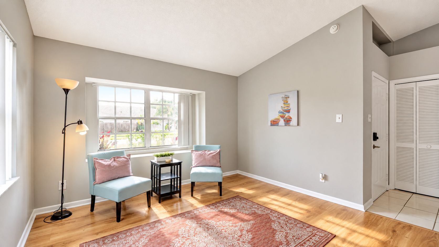 A room with a large window, two light blue armchairs, a small table with a plant, a floor lamp, a patterned rug, and a door with louvered shutters.