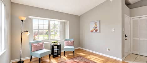 A room with a large window, two light blue armchairs, a small table with a plant, a floor lamp, a patterned rug, and a door with louvered shutters.