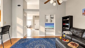 A living room with a leather sofa, a coffee table with breakfast items, a TV on a white stand, and a blue patterned rug.