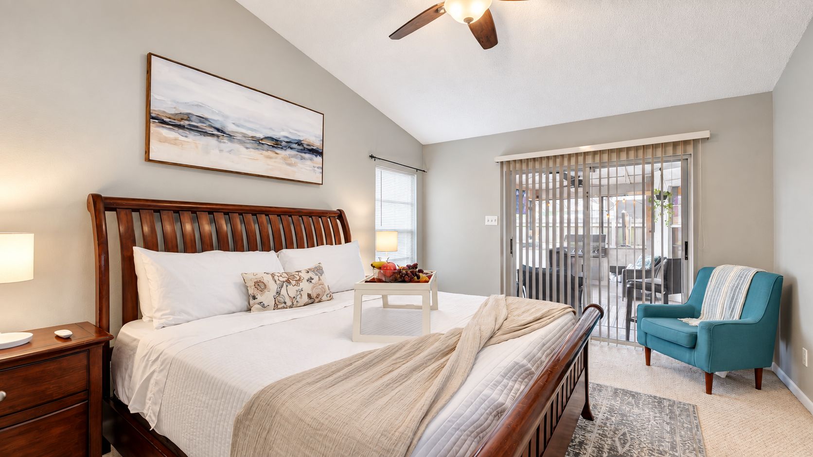 A bedroom with a bed, bedside table, lamp, and a view of the outside through a window with blinds.