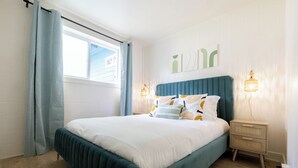 Una cama bien hecha con una cabecera color turquesa, ropa de cama blanca y almohadones decorativos. Hay dos lámparas de noche con un diseño tejido. La habitación tiene paredes blancas, una ventana con cortina y suelo de madera.