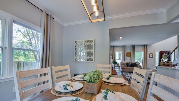 A dining area with a wooden table set for four, surrounded by white chairs. There is a woven basket with greenery on the table, and a decorative wall piece above it. - Bama Farmhouse - Beautiful Pool Area! (Tuscaloosa)