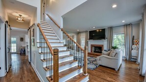 A spacious living room with a fireplace, a staircase, and a large TV mounted above the fireplace. - Bama Farmhouse - Beautiful Pool Area! (Tuscaloosa)