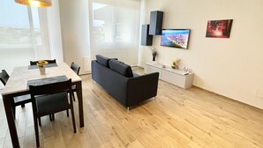 Una sala de estar moderna con suelo de madera, un sofá, una mesa de comedor con sillas, un televisor montado en la pared y un ventanal con vistas.