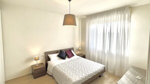 Un dormitorio ordenado con cama, lámparas de noche, mesita de noche y ventana con cortinas traslúcidas.