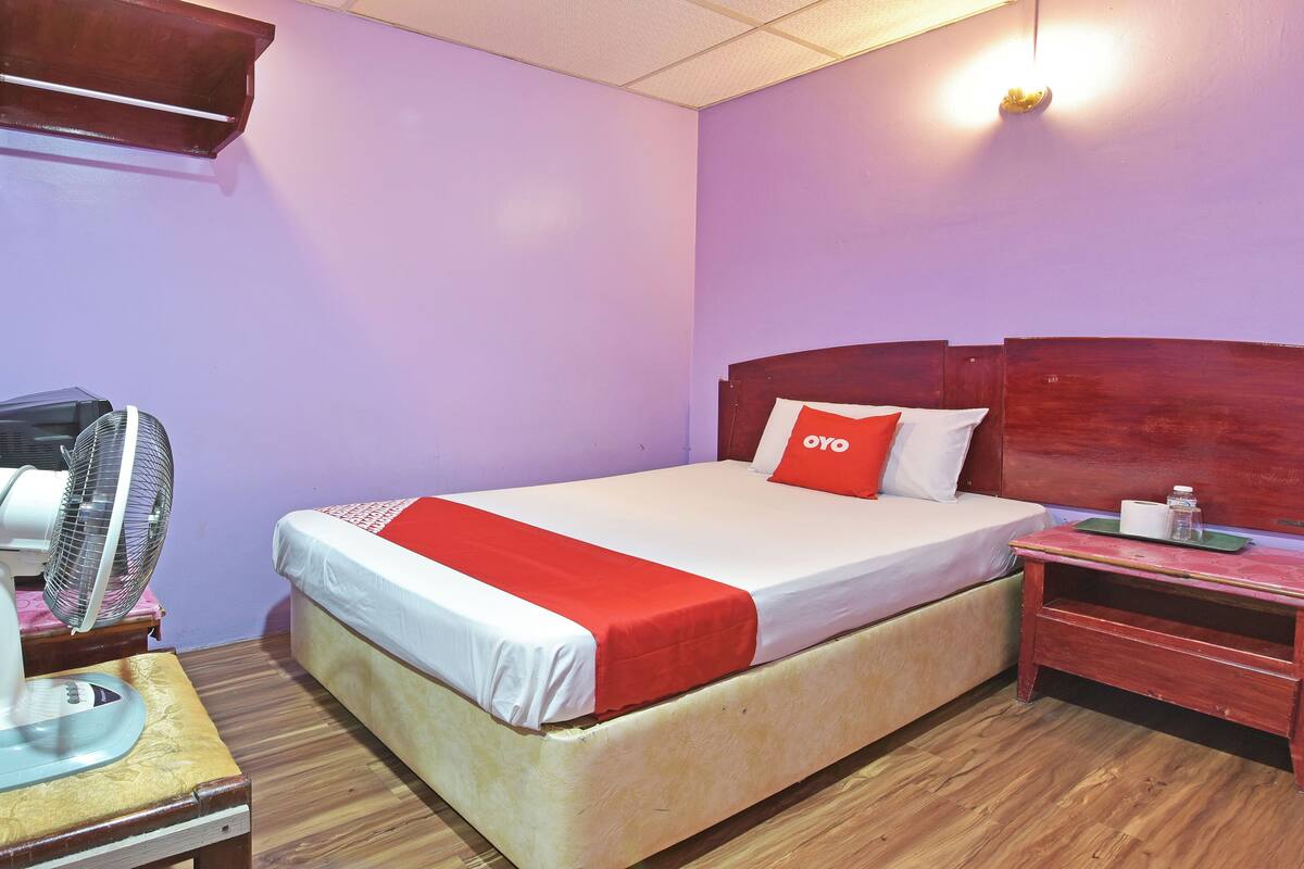 A single bed with a wooden headboard, a bedside table with a lamp, and a wall-mounted fan.