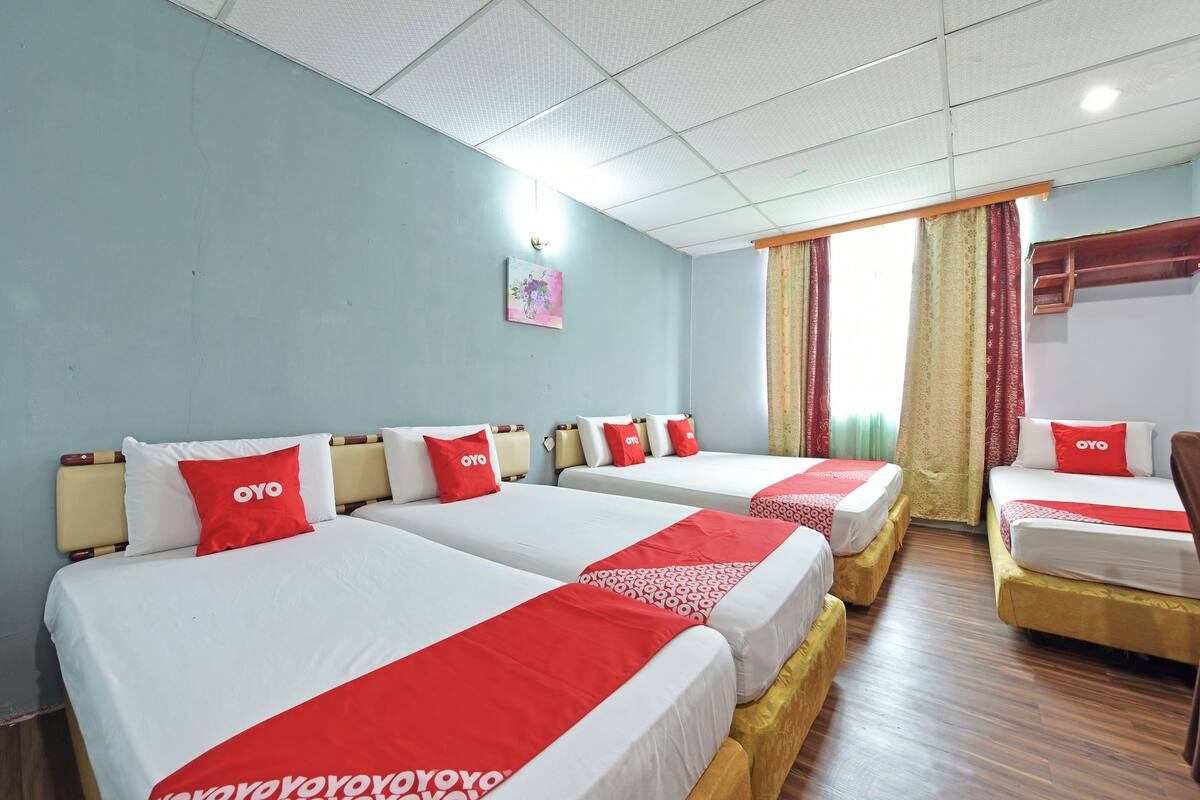 A hotel room with three beds, each with a red and white bedspread and pillows labeled "OYO". The room has light blue walls, a wooden floor, and a window with curtains.