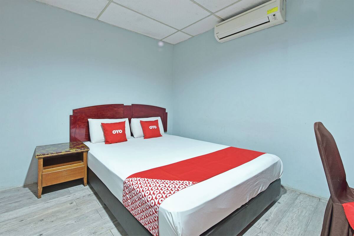 A neatly made bed with a red and white bedspread, a wooden nightstand with a drawer, and a brown chair.