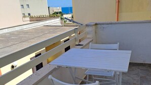 A rooftop terrace with a white table and two white chairs, a metal railing, and a view of the sea and buildings in the background. - Olga's House  (Otranto)