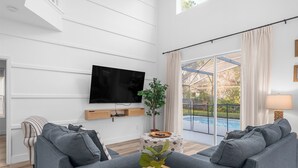 A modern living room with a flat-screen TV, grey sofas, a wooden coffee table, and a large window with a view of a swimming pool. - Movie & Game rooms/Car Charger/Pool (Kissimee)